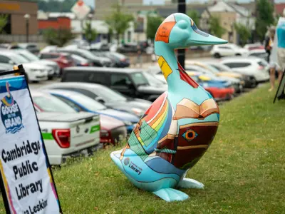 A large, hand-painted Cambridge Goose sculpture featuring books, rainbow colours, and literary references stands on grass beside a Goose on the Grand event sign for the Cambridge Public Library.