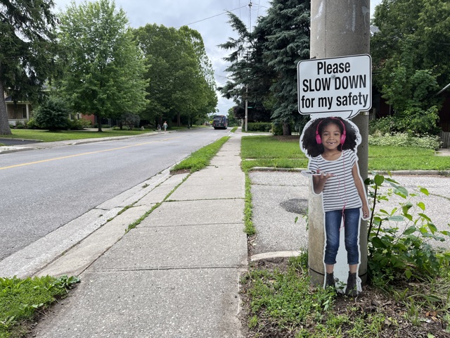 A poster with a small child that has headphones on by the road. Textbox says "Please SLOW DOWN for my safety"