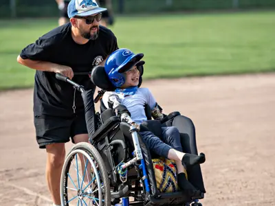 Participant wearing a blue batting helmet sits in a specialized sports wheelchair on a baseball diamond while another participant pushes from behind. The pair move across the infield during a Buddy League baseball game, with a grassy outfield and fence visible in the background.