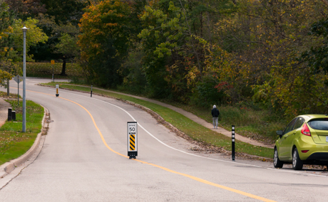 A two‑lane road in a wooded residential area with several traffic-calming signs and posts placed along the centreline. A speed limit sign reading “50” is mounted on one of the centreline devices. A green car is parked on the right side of the road, and a person walks along the sidewalk in the background.
