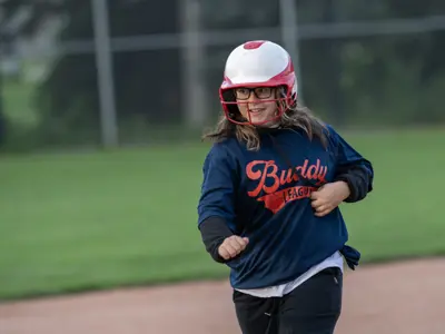 Participant wearing a red-and-white batting helmet and a Buddy League jersey runs along the baseball diamond during a game. The photo captures the participant in motion with the field and outfield fence softly blurred in the background.