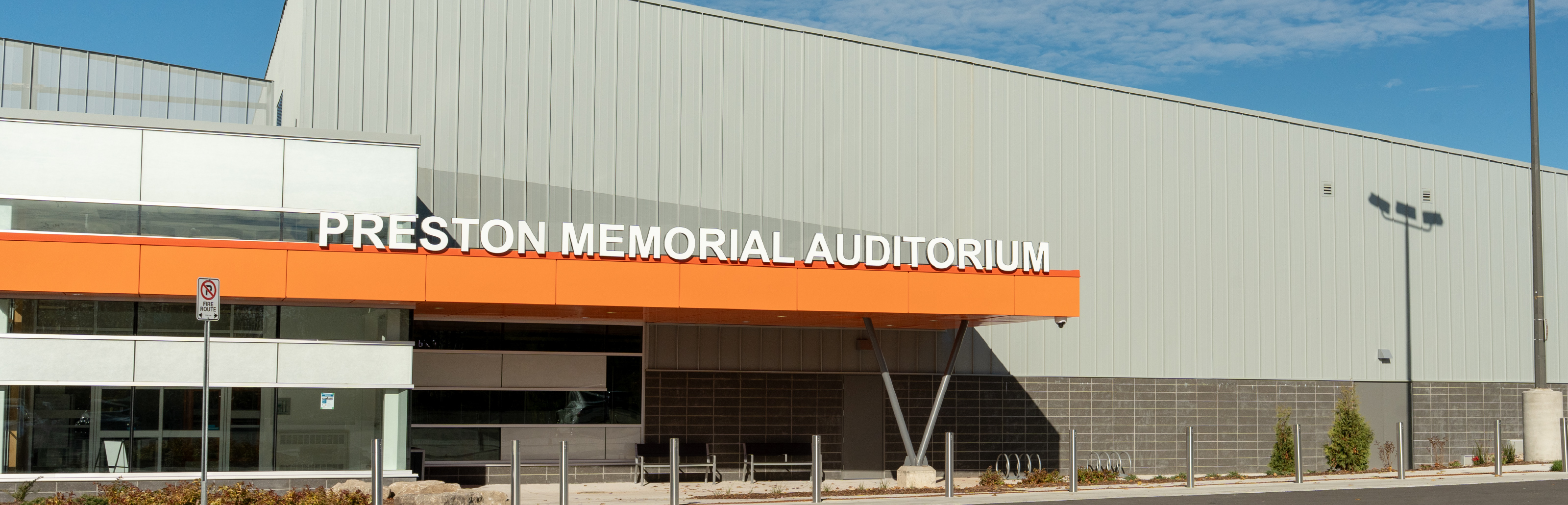 Exterior view of the Preston Memorial Auditorium building on a clear day. Large white letters spelling “PRESTON MEMORIAL AUDITORIUM” are mounted above a bright orange entrance canopy. The modern facility features glass windows, grey metal cladding, a paved walkway, bike racks, and landscaping in front of the entrance.