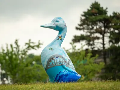 A large, hand-painted Cambridge Goose sculpture in shades of blue with butterflies and a bridge design stands on a grassy hill at the Goose on the Grand event.