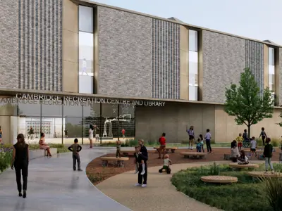 Architectural rendering of the future Cambridge Recreation Complex. The modern building features a brick and glass exterior surrounded by landscaped gardens, trees, and pedestrian pathways. Community members gather throughout an outdoor plaza with seating, play areas, and green spaces leading to the main entrance.