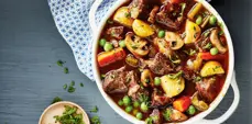 A white oval casserole dish filled with hearty beef stew, featuring chunks of beef, potatoes, mushrooms, carrots, peas, and herbs in a rich broth, viewed from above.