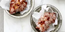 Two glazed apple fritters sit on parchment paper in rustic ceramic bowls on a light marble surface, photographed from above.