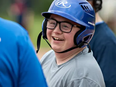 Close-up of a Buddy League baseball participant wearing a blue batting helmet and glasses. The participant stands near teammates on the field, with the baseball diamond and players visible in the background.