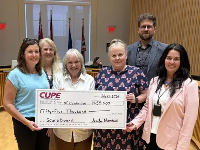 Six people stand in Council Chambers holding an oversized cheque from CUPE Local 1882 to the City of Cambridge for $55,000 in support of a scoreboard project. Behind the group are council seating areas and flags displayed at the front of the chamber.