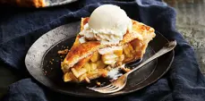 A slice of golden apple pie topped with a scoop of vanilla ice cream rests on a dark plate beside a fork, with a navy cloth backdrop.