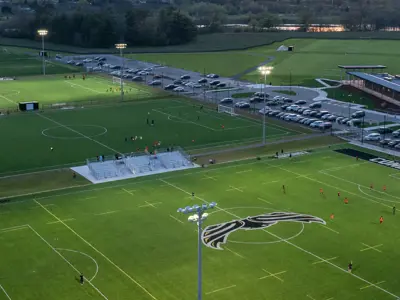 Aerial view of the Cambridge Soccer Complex at dusk, showing several illuminated soccer fields with players in action. The facility includes spectator seating, a clubhouse, parking areas, and walking paths, surrounded by open green space and nearby roadways.