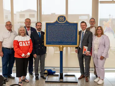 Seven people stand on either side of a blue and gold Ontario Heritage Trust plaque for the Galt Football Club inside a building with large windows.