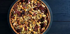 Overhead view of a caramelized honey nut tart topped with pecans, hazelnuts, sliced almonds, pumpkin seeds, and sunflower seeds on a dark blue wooden surface.