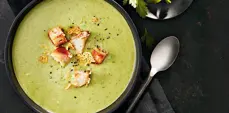 Creamy green broccoli soup served in a dark bowl, topped with golden croutons and cracked black pepper, with a spoon resting beside the bowl on a dark tabletop.