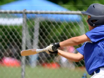 Action photo of a youth baseball player in a blue uniform and black batting helmet swinging a bat at an incoming baseball. The player stands in front of a chain-link fence on an outdoor baseball field, with blurred spectators, tents and greenery visible in the background. The image captures the ball approaching the bat during play.